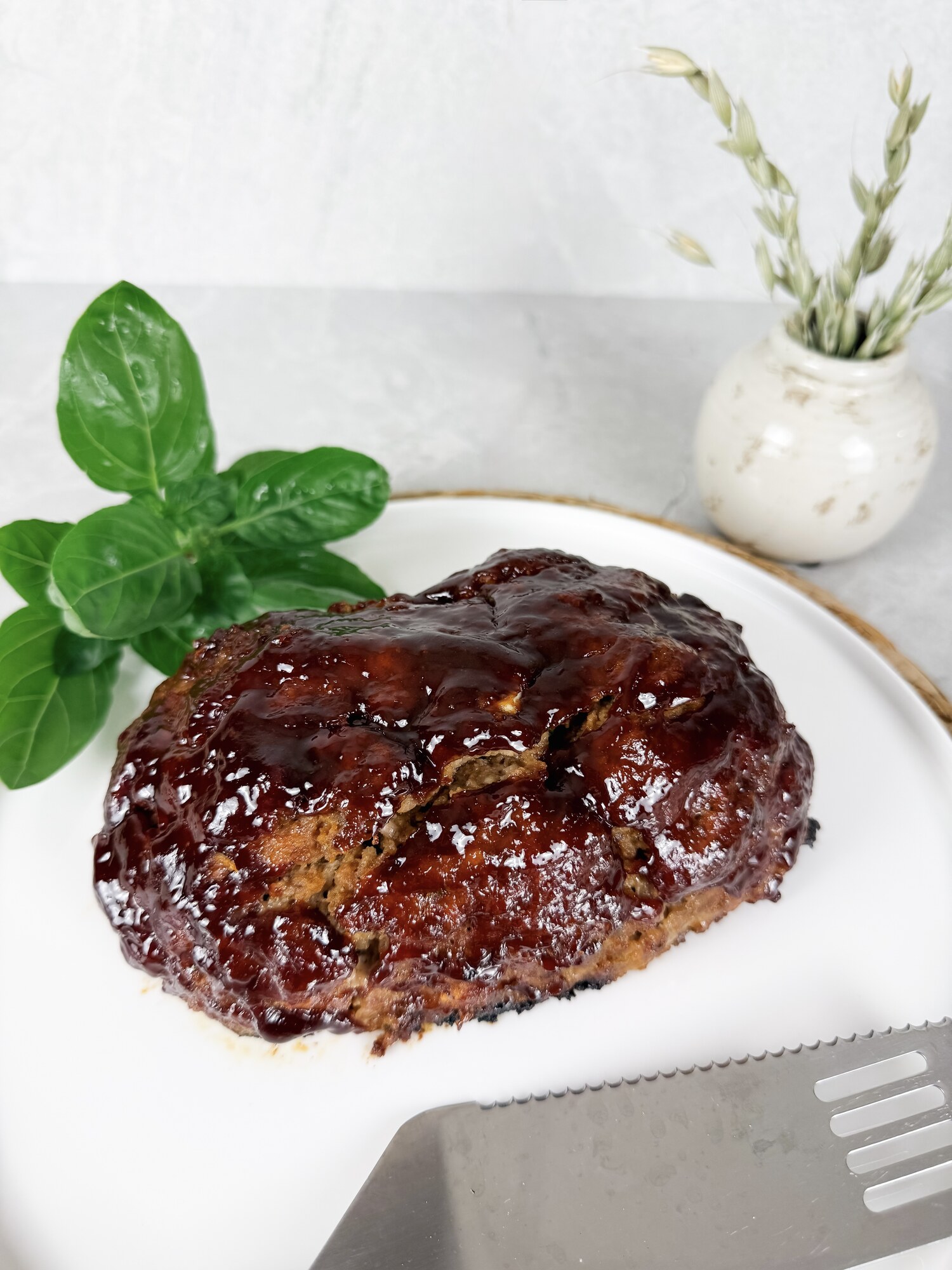 A whole balsamic-glazed meatloaf on a white plate with fresh herbs and a small vase of dried oats