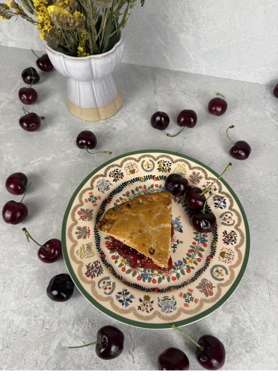 A slice of cherry pie on a painted plate, fresh cherries scattered around