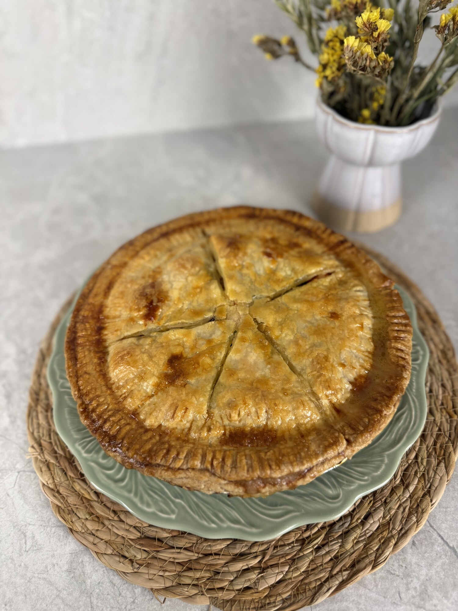 A golden chicken potpie on a green plate, with a small vase of dried yellow flowers behind it