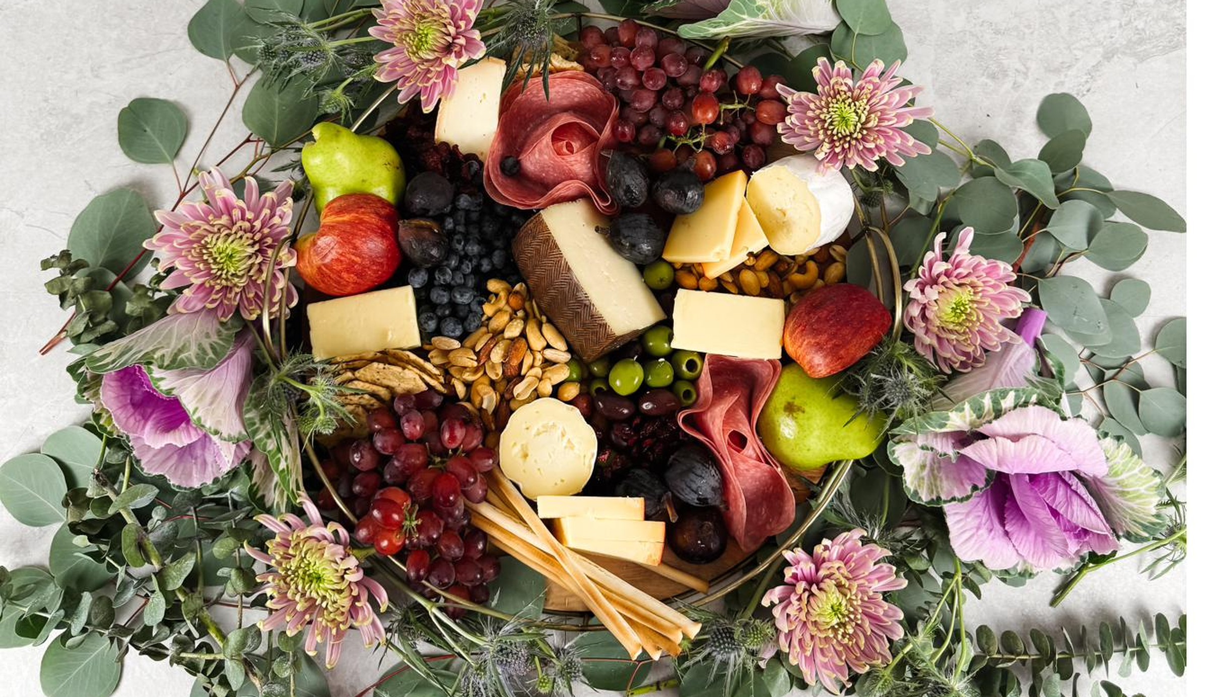 A charcuterie board with cheeses, fruit, and flowers