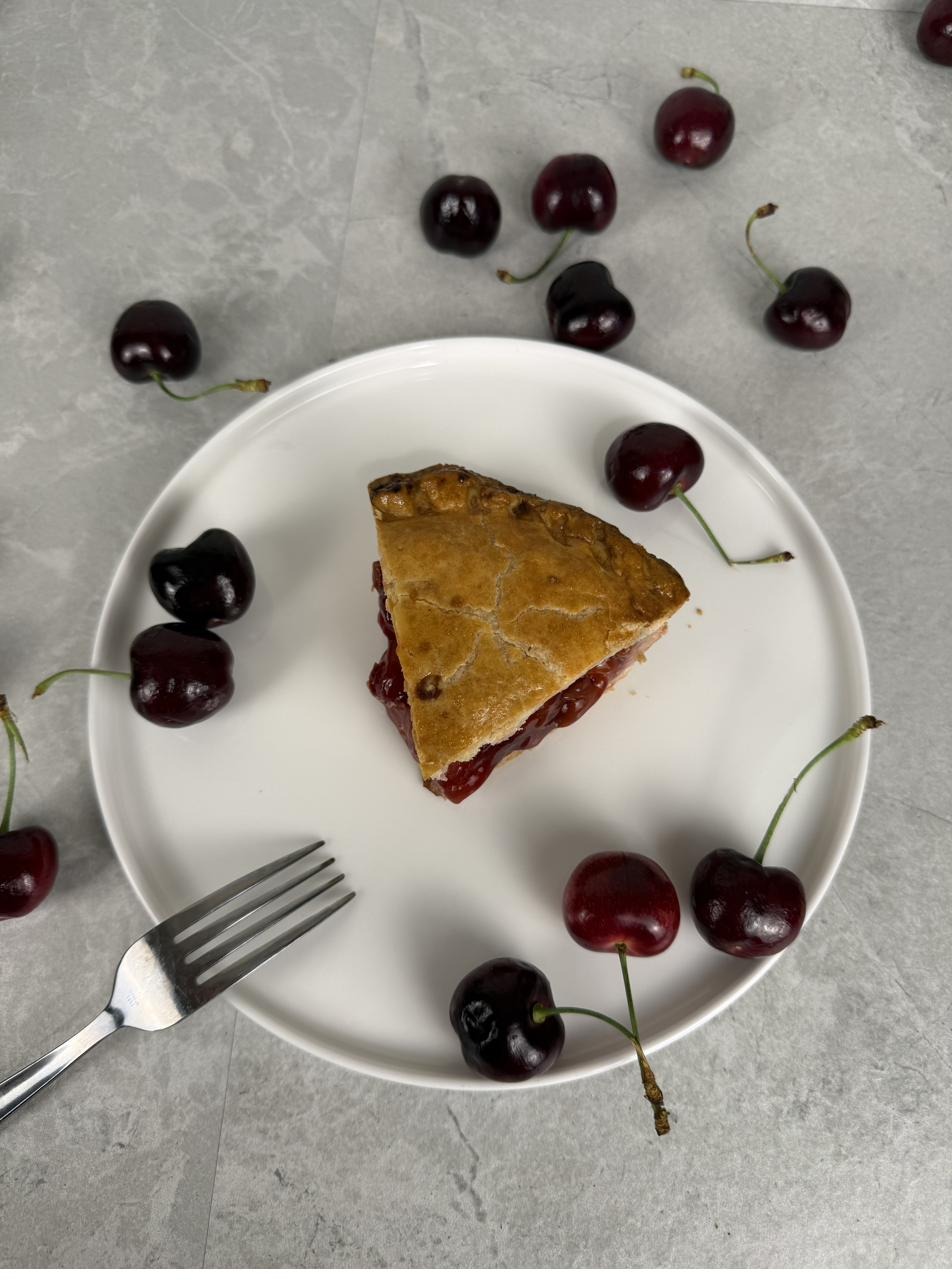 A slice of cherry pie on a white plate with fresh cherries