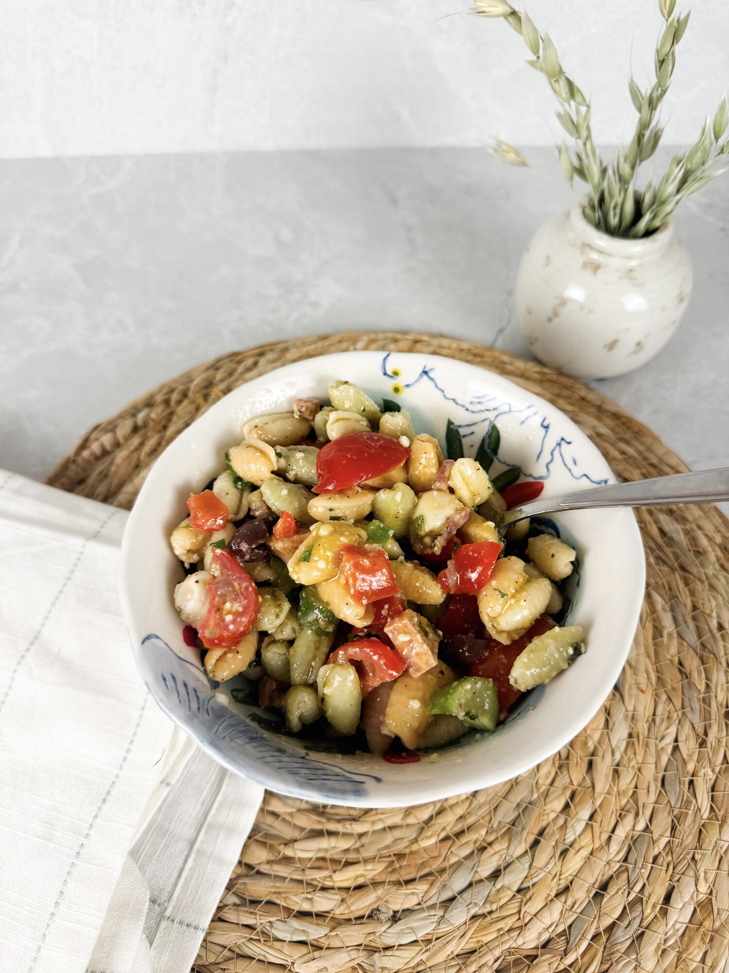 A blue-and-white bowl of antipasto pasta salad on a woven mat, with a linen napkin and a vase of dried oats