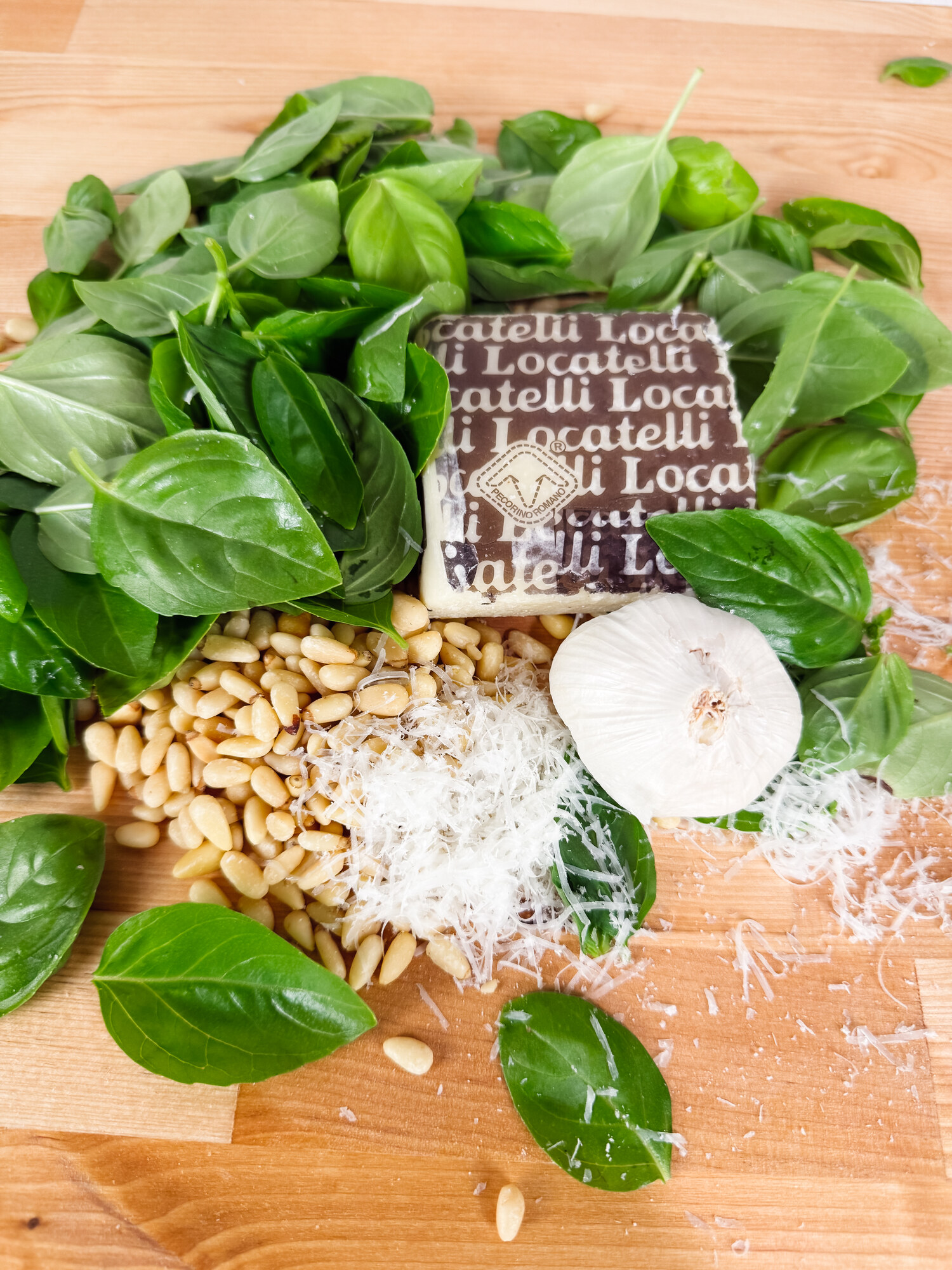 Fresh basil, toasted pine nuts, a wedge of Locatelli Pecorino Romano, grated cheese, and garlic on a wooden board