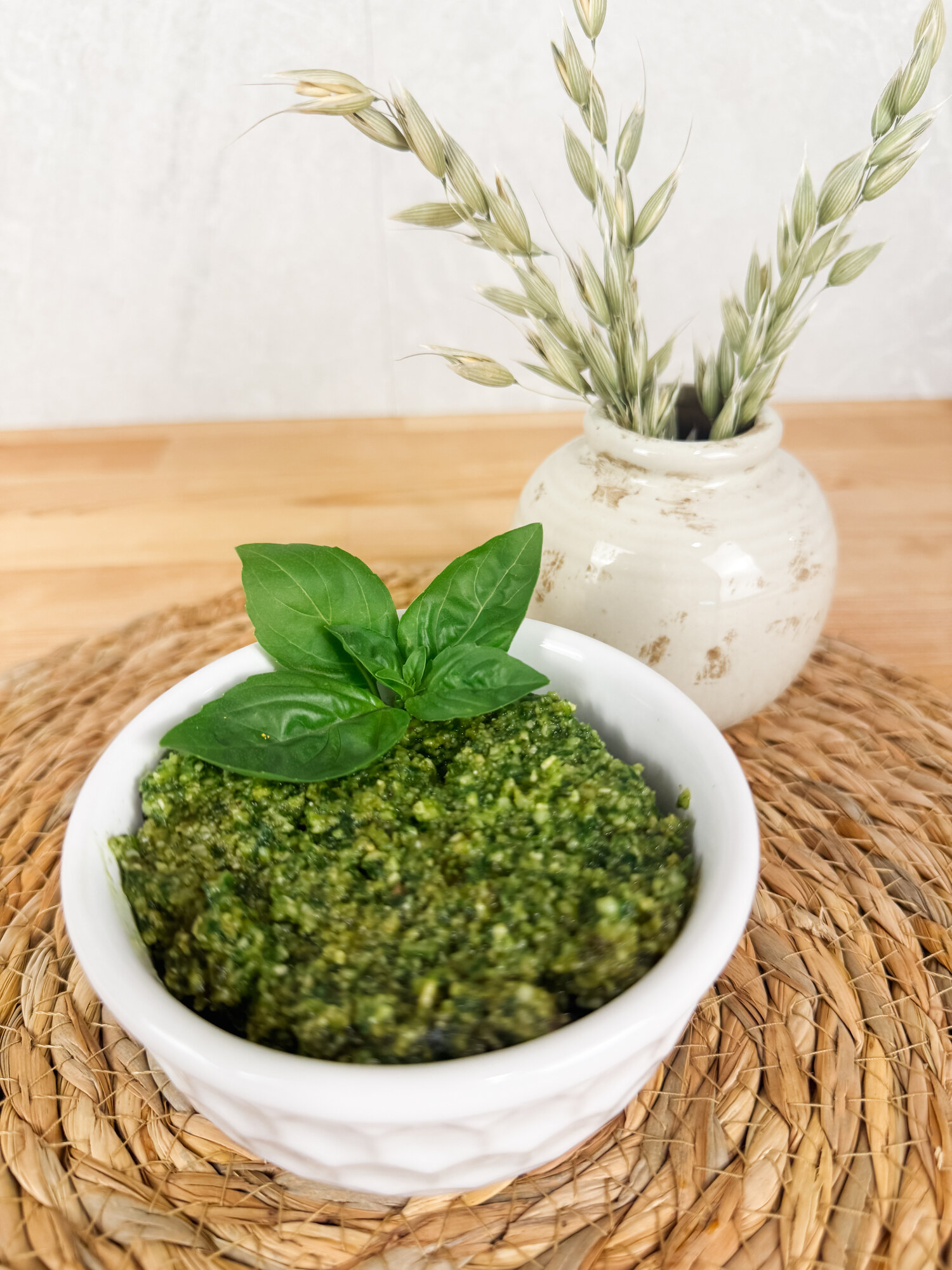 A white bowl of fresh basil pesto topped with a basil sprig, on a woven mat with a vase of dried oats behind